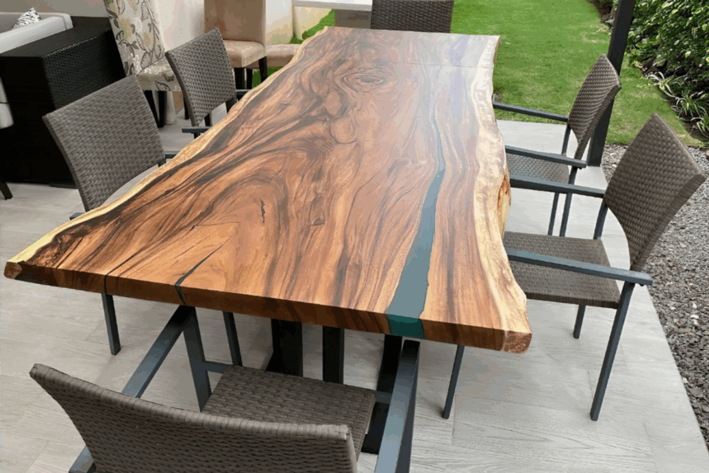 Wood table w/ natural edges & resin river, surrounded by 6 woven chairs on patio.