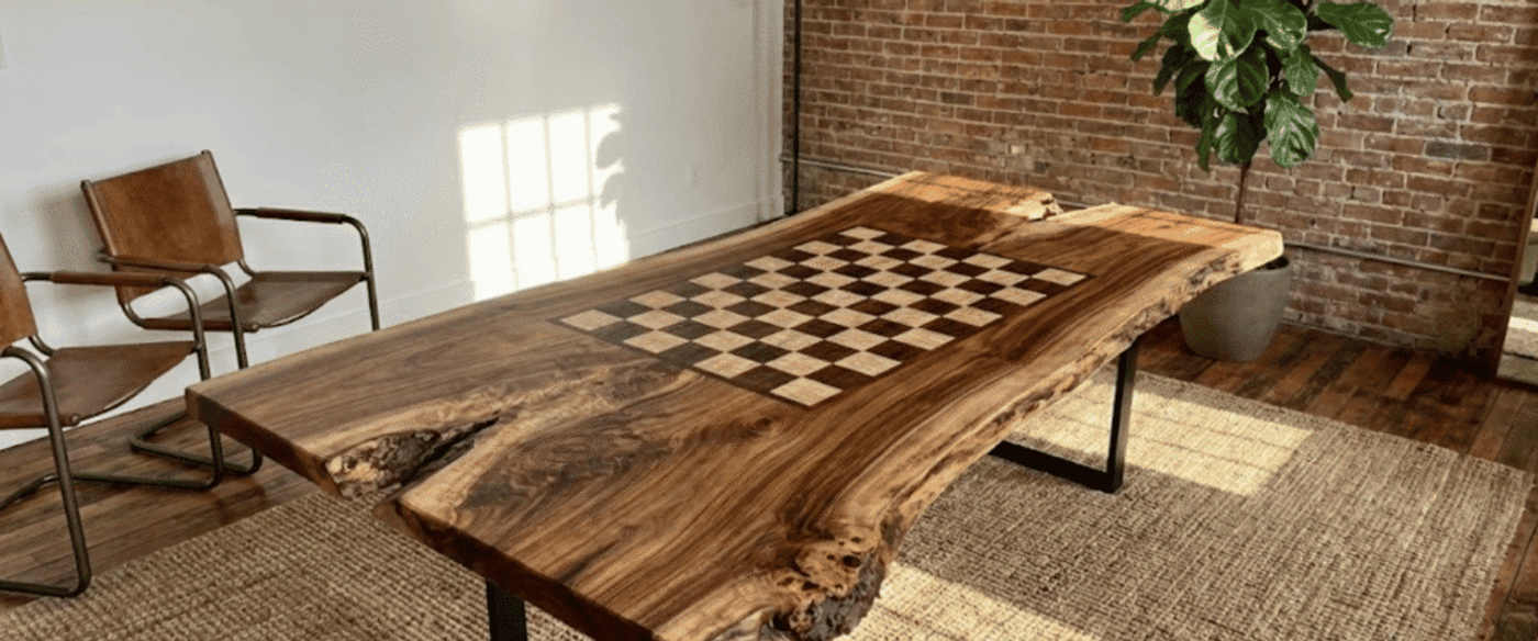 Live-edge walnut table with inlaid chessboard center, industrial metal legs, leather chairs, brick wall and potted fiddle-leaf fig plant