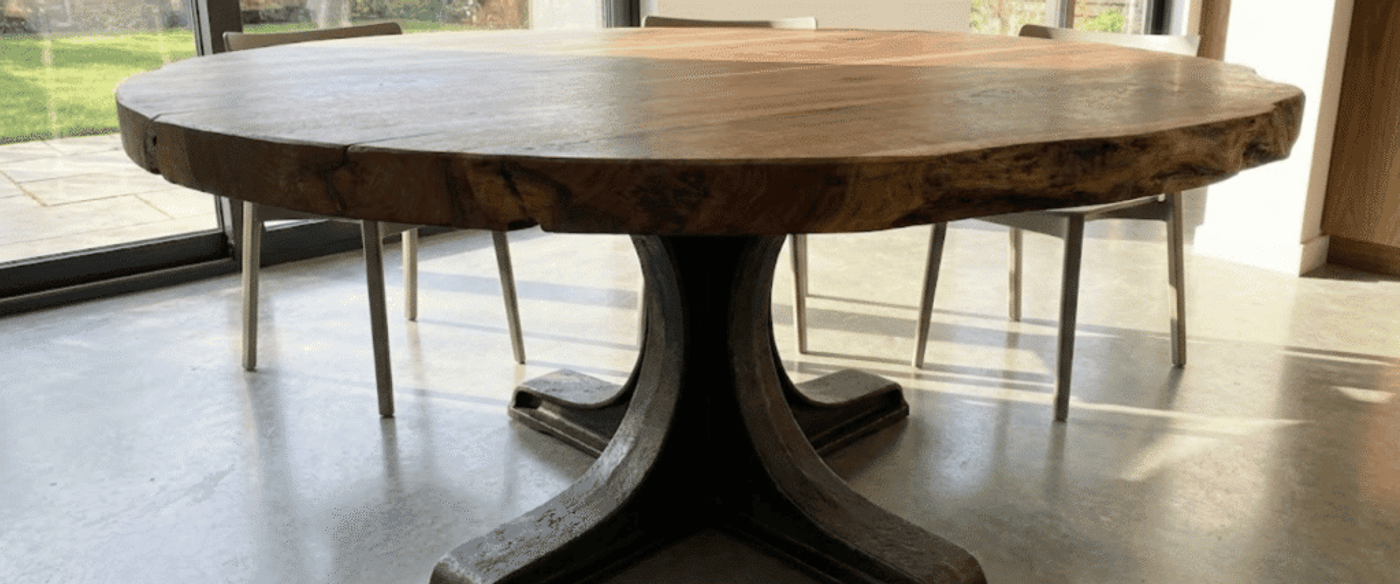 Round live-edge wooden dining table with a sturdy black cast-iron pedestal base, chairs and sunlit glass doors in background