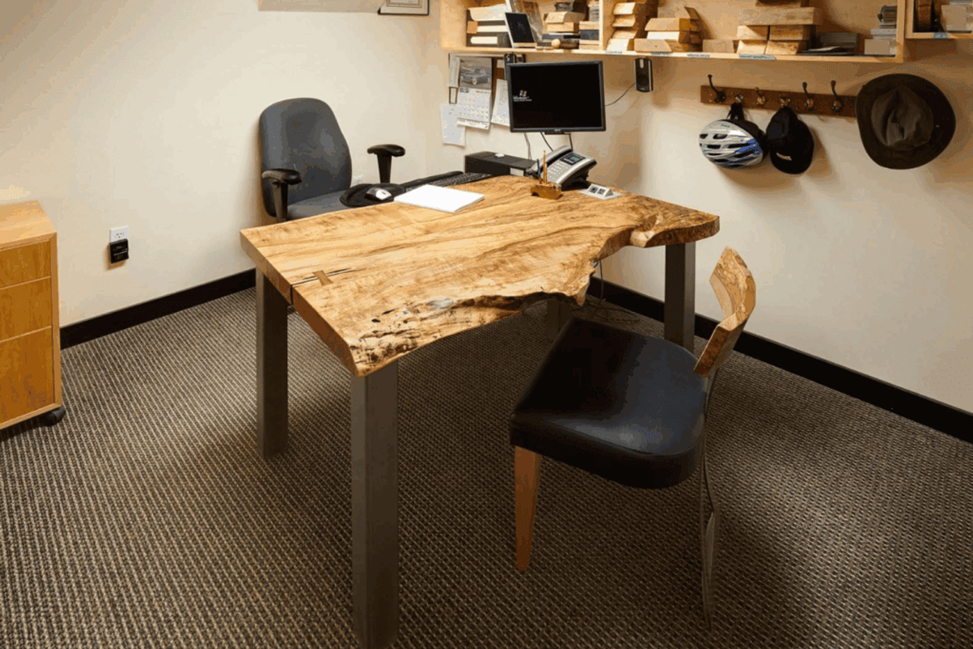 Wooden office desk with natural edges, two chairs, computer setup, and shelves with wooden blocks and hats.