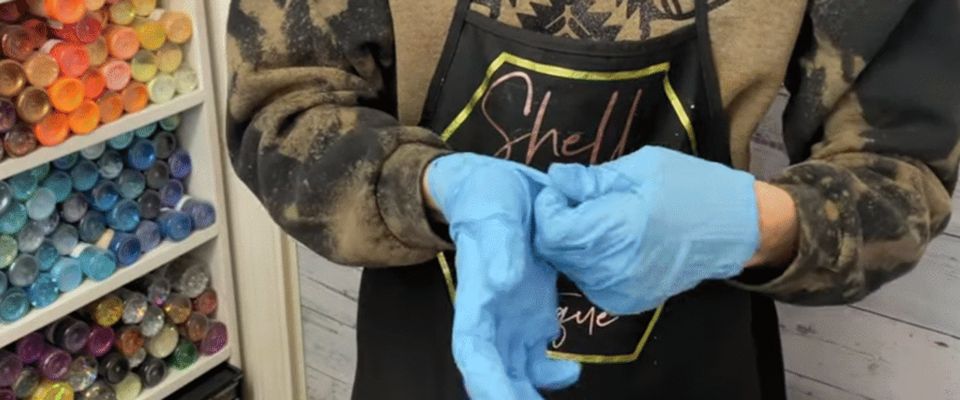 Person wearing an apron putting on blue disposable gloves, with shelves of colorful paint or pigment containers visible in the background.