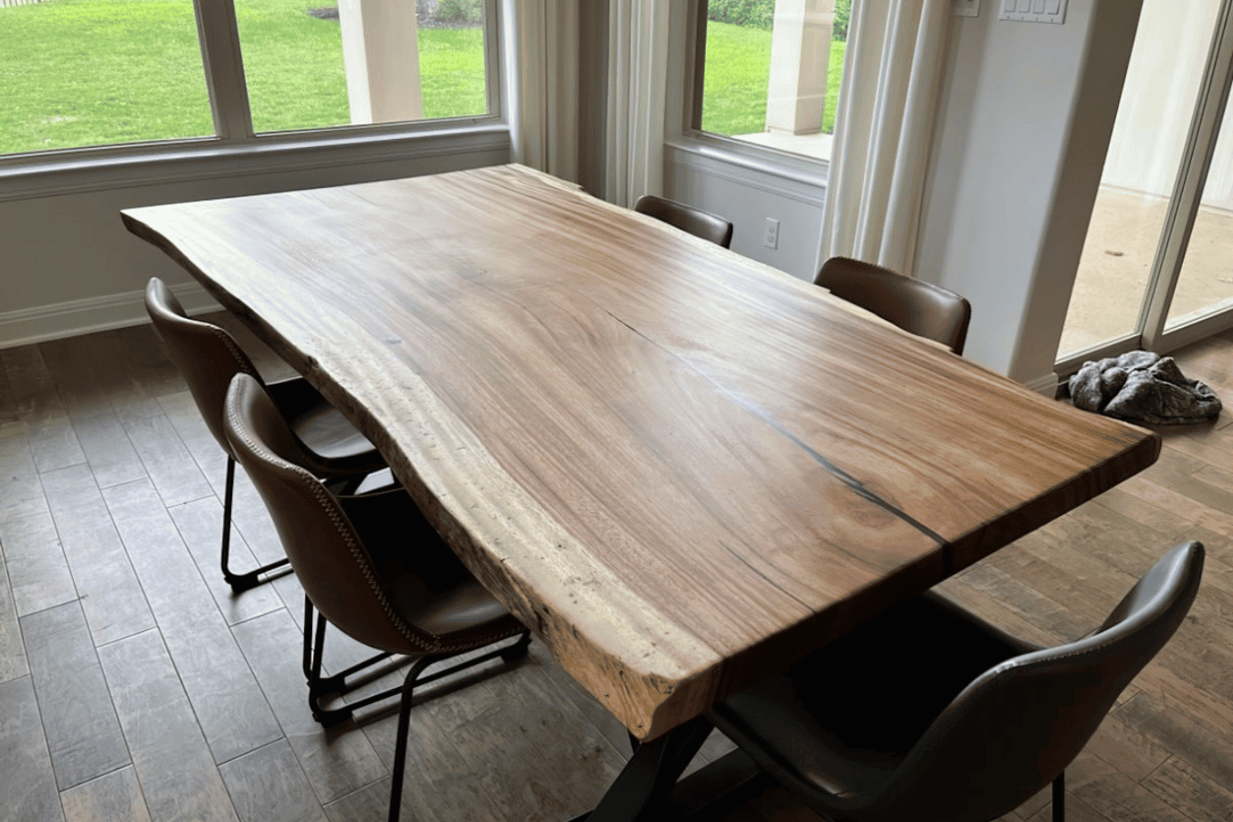 Live-edge walnut dining table with six leather chairs in a sunlit dining room, hardwood floors and large windows overlooking green lawn.