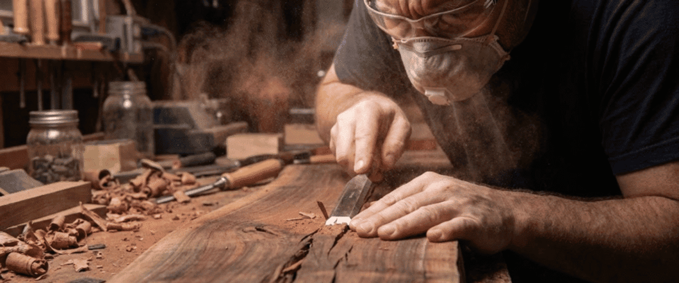 Woodworker wearing mask and glasses carefully chisels a live-edge slab, surrounded by wood shavings and hand tools