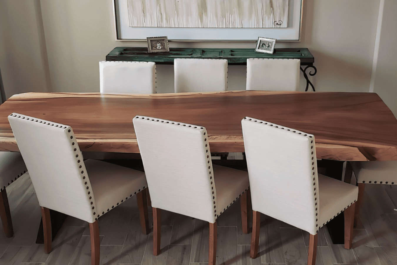 Dining room with a natural wood slab table and six beige upholstered chairs, set on a wooden floor. Background features a green console table with framed photos and a modern artwork on the wall.