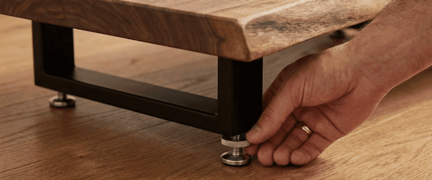 Hand adjusting metal leveling foot on a black steel leg supporting a live-edge wooden table on a hardwood floor