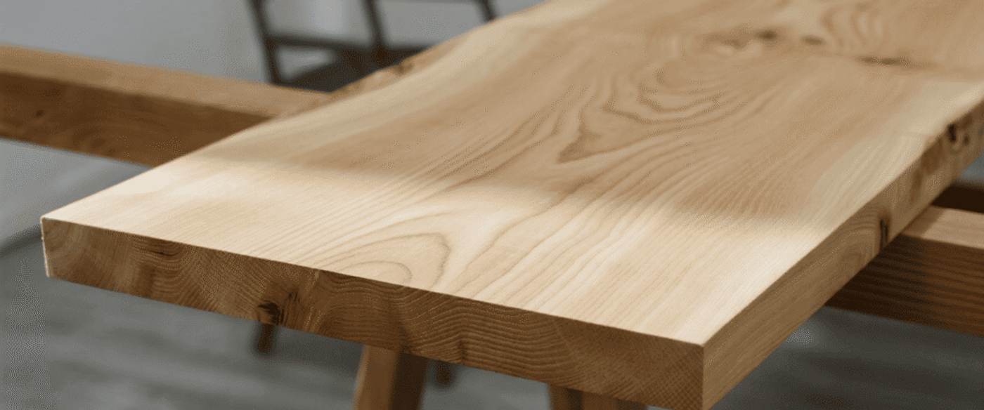 Smooth unfinished wooden plank with visible grain resting on sawhorses in a workshop, soft light and blurred chair in background.
