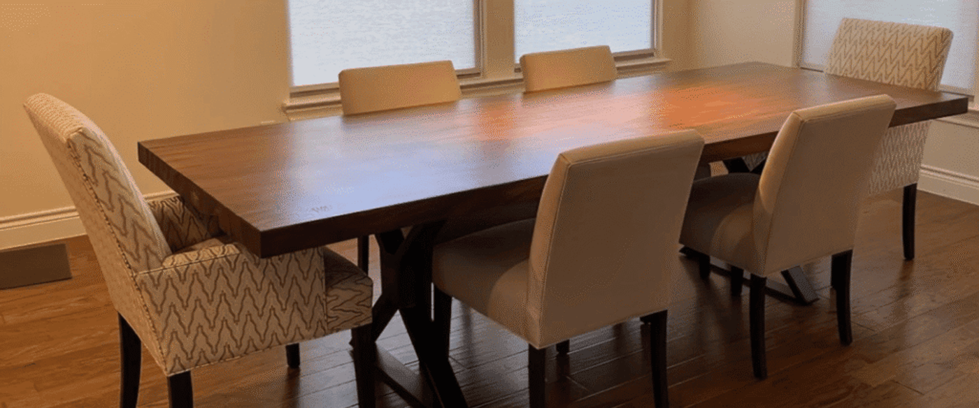 Dining room with long wooden table and six upholstered chairs, three windows with shades, warm hardwood floor, minimal decor.