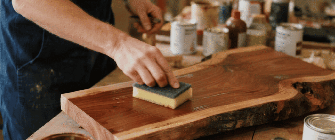 Applying wood finish with a sponge to enhance grain and protect the surface of a natural wooden board in a workshop.