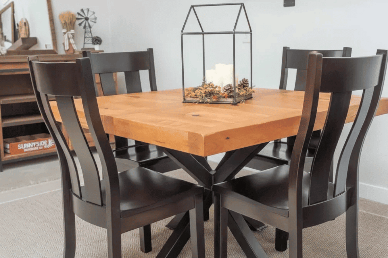 Wooden square dining table with black pedestal base surrounded by four dark wood chairs, candle centerpiece in glass terrarium.