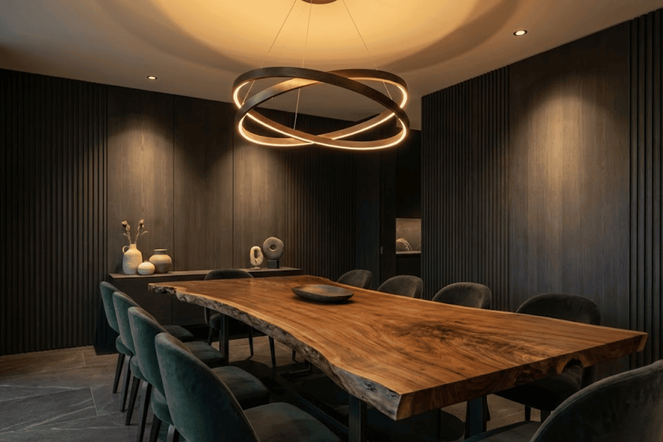 Dining room with a live-edge wooden table, green velvet chairs, sculptural chandelier, and dark textured walls, warm mood lighting