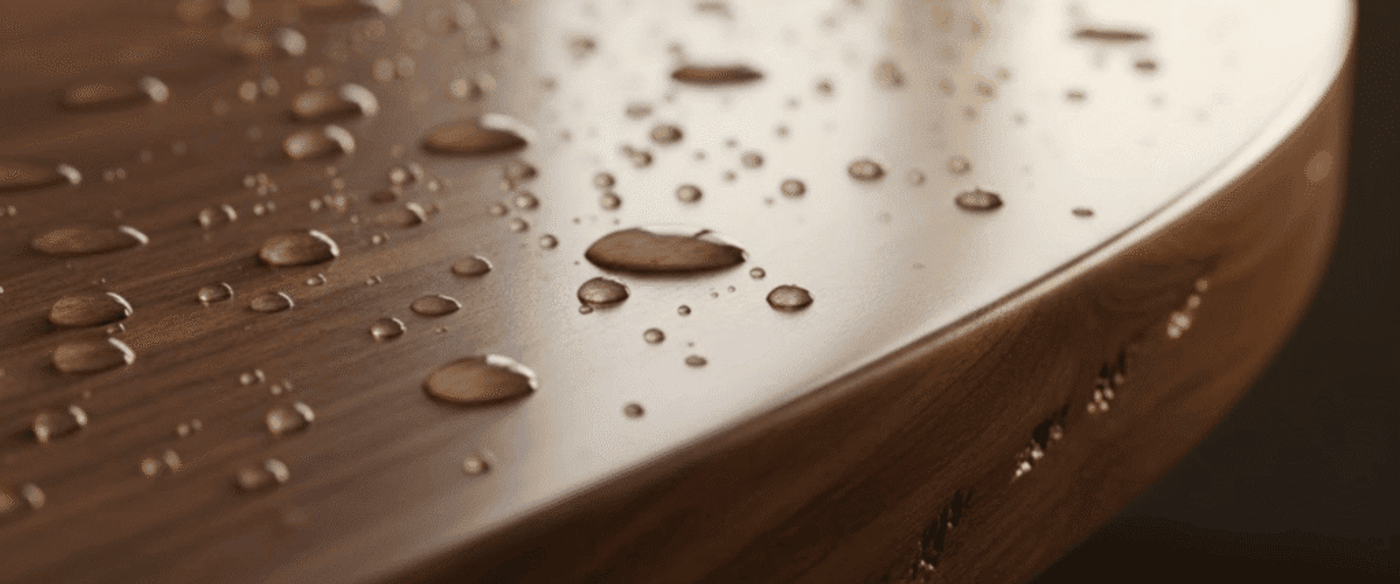 Round wooden table edge with scattered water droplets reflecting soft window light, highlighting wood grain and smooth finish