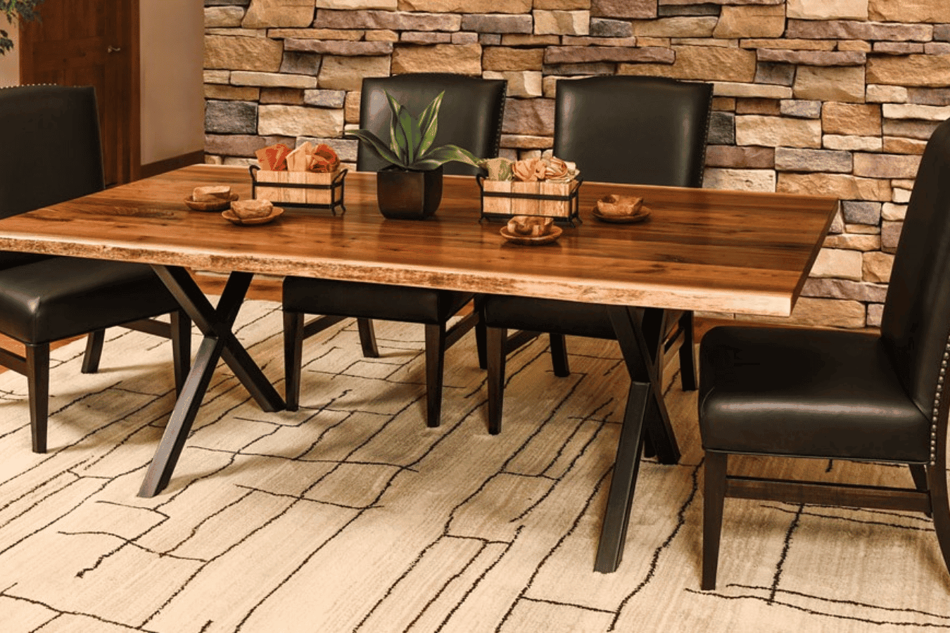 Wood dining table with black leather chairs on a patterned rug, stone accent wall, decorative bowls and a potted green plant.