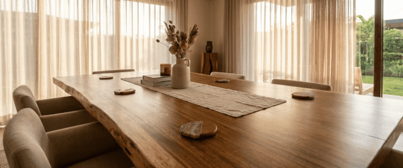 Warm, sunlit dining room with a large live-edge wooden table, beige upholstered chairs, vase of dried flowers, and sheer curtains
