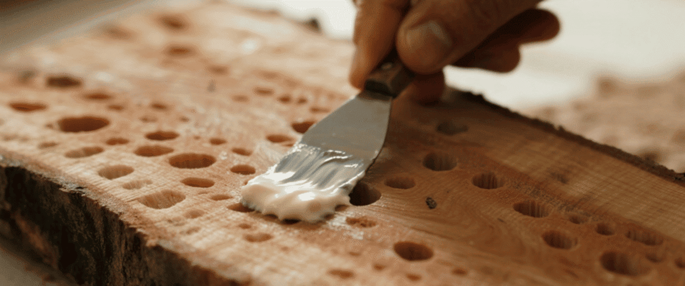Hand spreading wood filler into drilled holes on a wooden plank with a small putty knife.