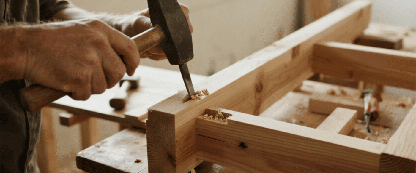 Carpenter chiseling a mortise joint in a wooden beam on a workbench, wood shavings scattered nearby.
