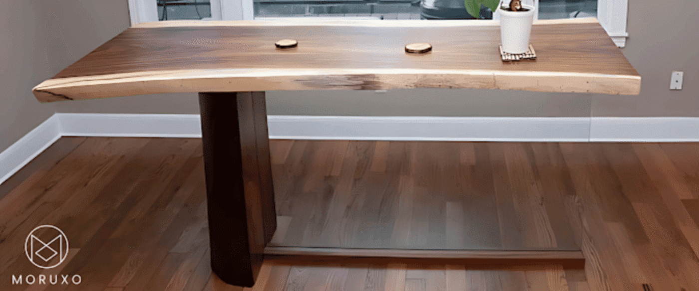 Live-edge walnut dining table with two round wooden coasters and potted plant on polished hardwood floor near window, modern single-leg base.