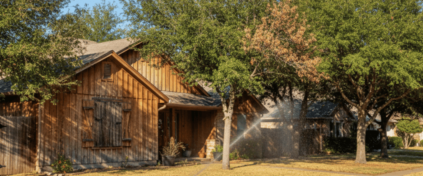 Sunlit wooden suburban house with lawn sprinklers watering grass, shaded by mature trees