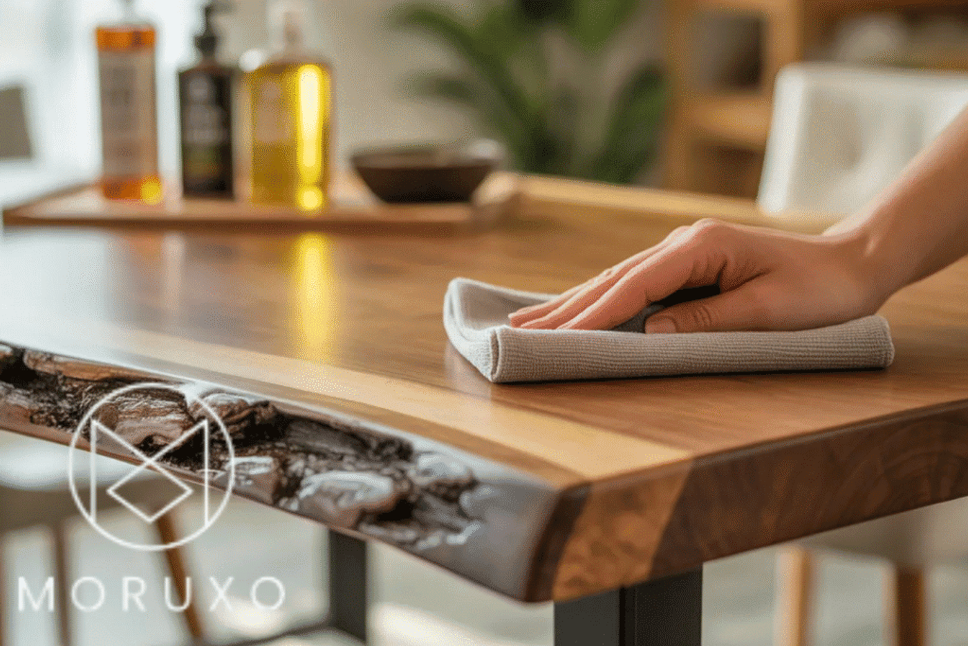 Hand wiping a polished wooden table with a cloth, showing natural wood grain and live edge detail. Bottles blurred in background
