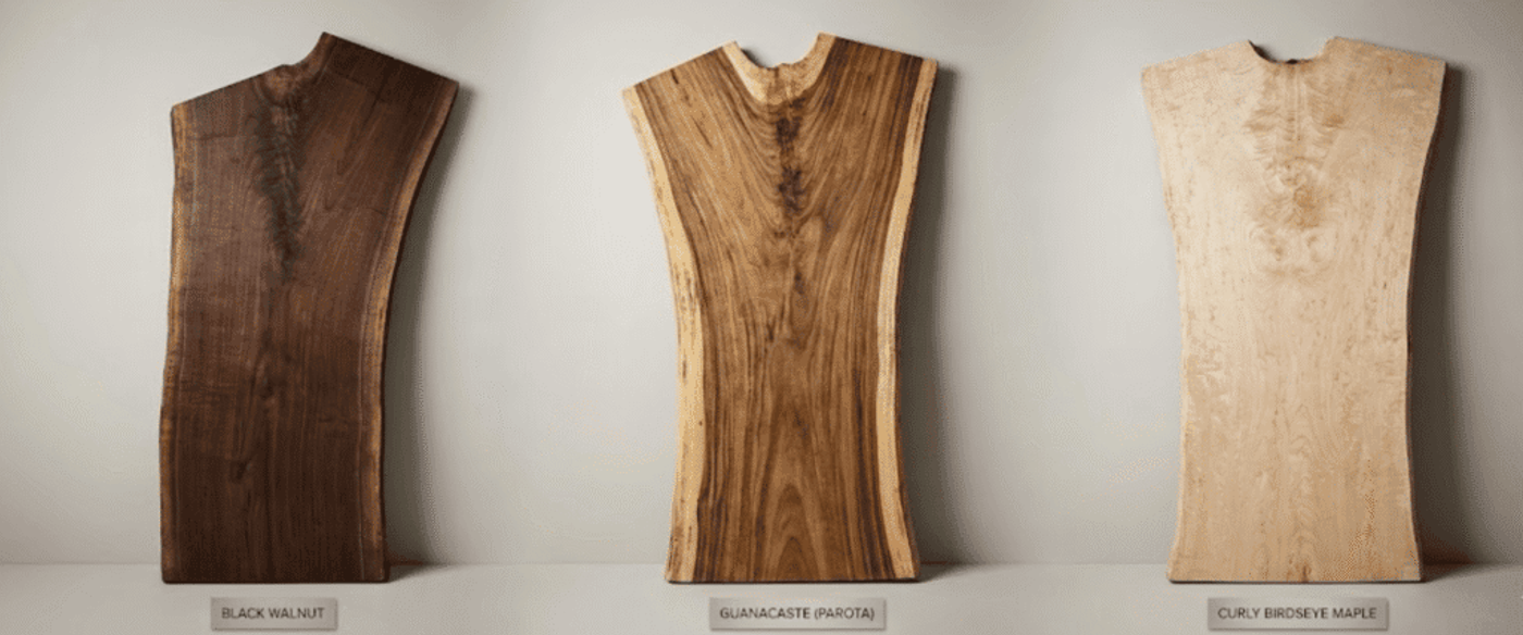 Three live-edge wood slabs (black walnut, guanacaste, curly birdseye maple) standing upright against a neutral wall