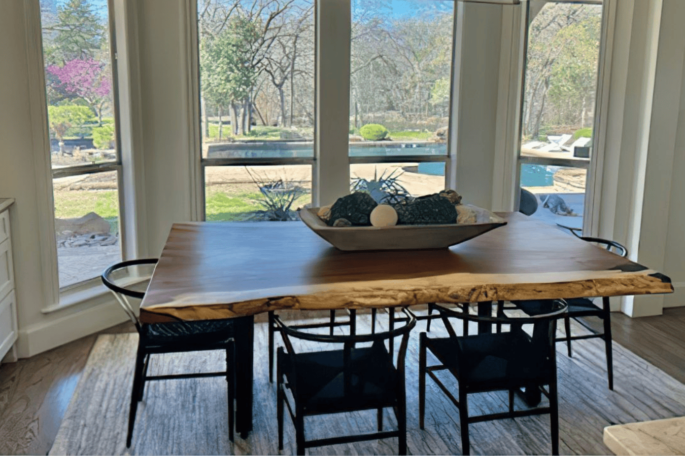 Dining table with live-edge wood top and black chairs, positioned by large windows overlooking a garden and pool.