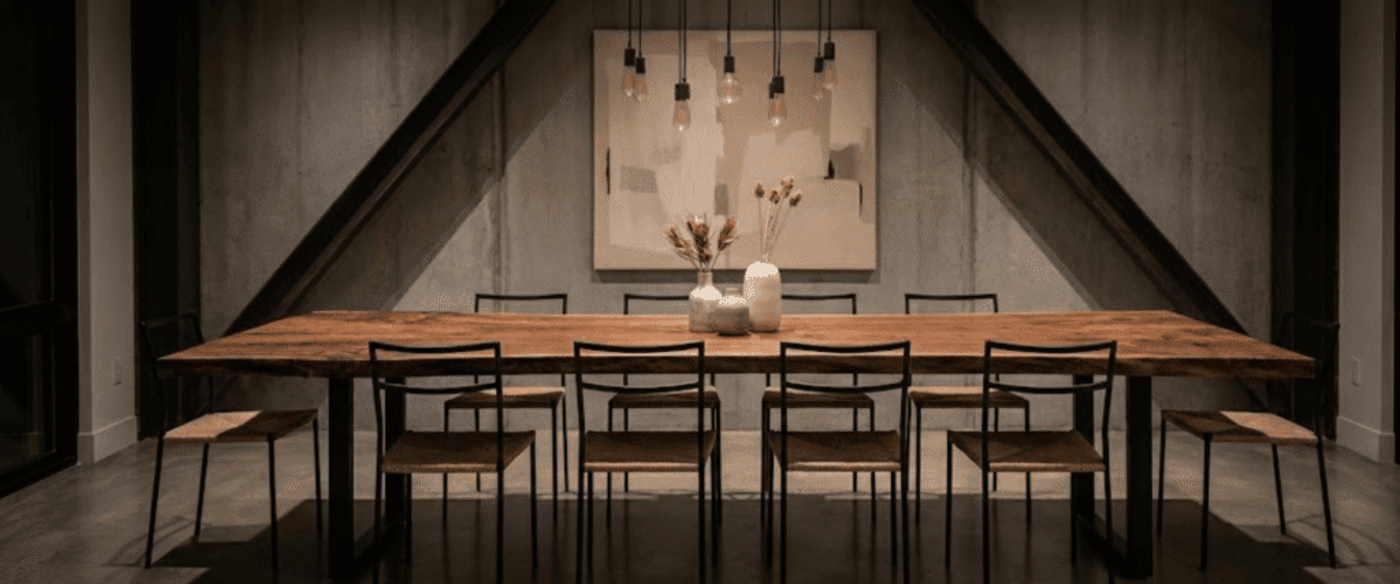 Long wooden dining table with eight simple chairs under hanging bulbs, minimalist concrete walls and vase centerpiece, dim light