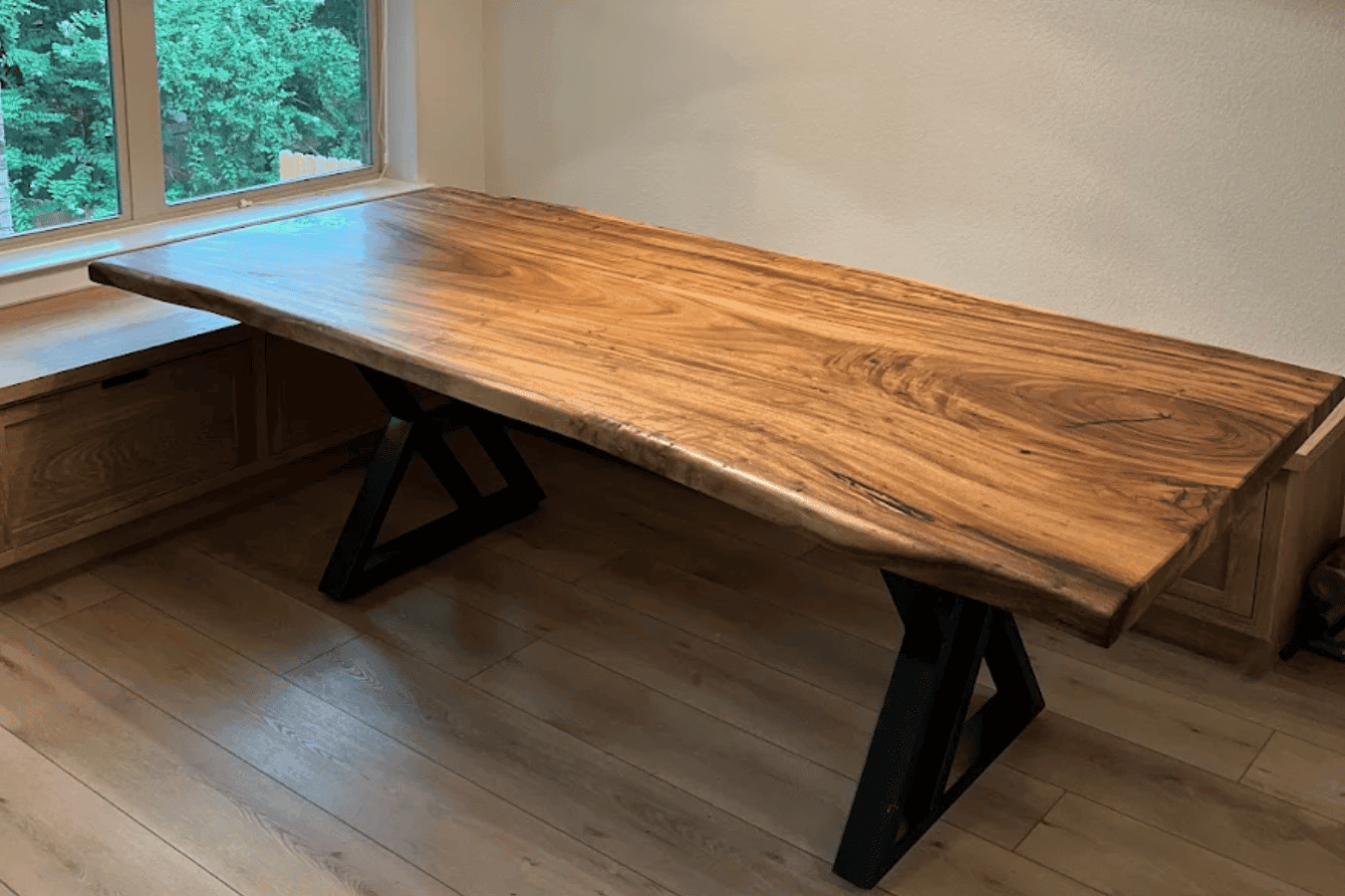 Solid live-edge wooden dining table with black metal X-legs under a woven pendant light, next to a window seat and large window showing green trees.