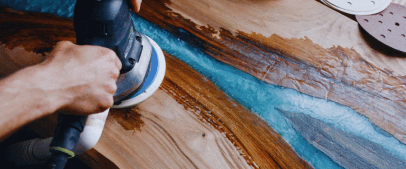 Hand polishing wooden surface with blue epoxy resin using an electric sander.