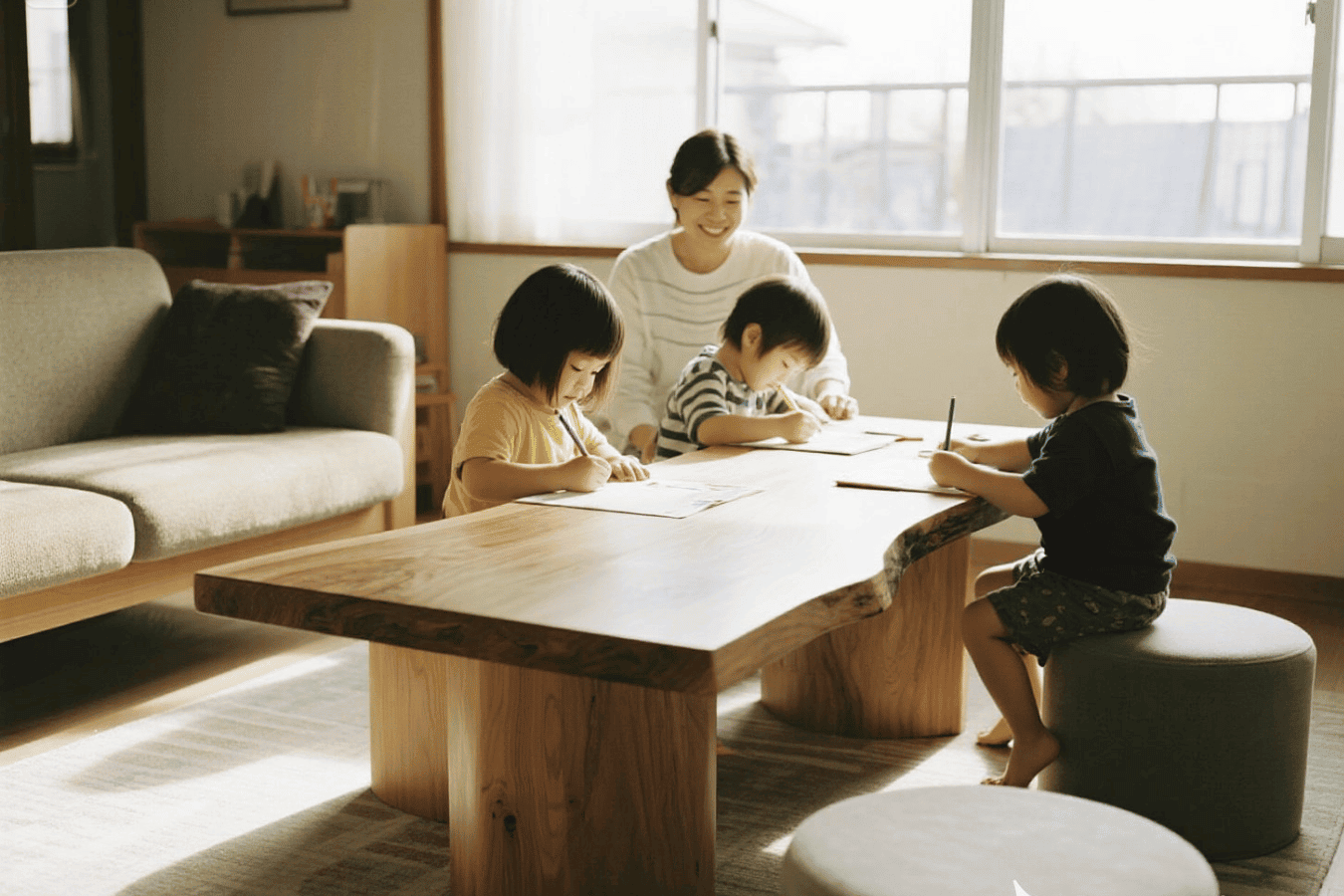 Three children and an adult seated around a low wooden table, drawing and writing together in a sunlit living room.