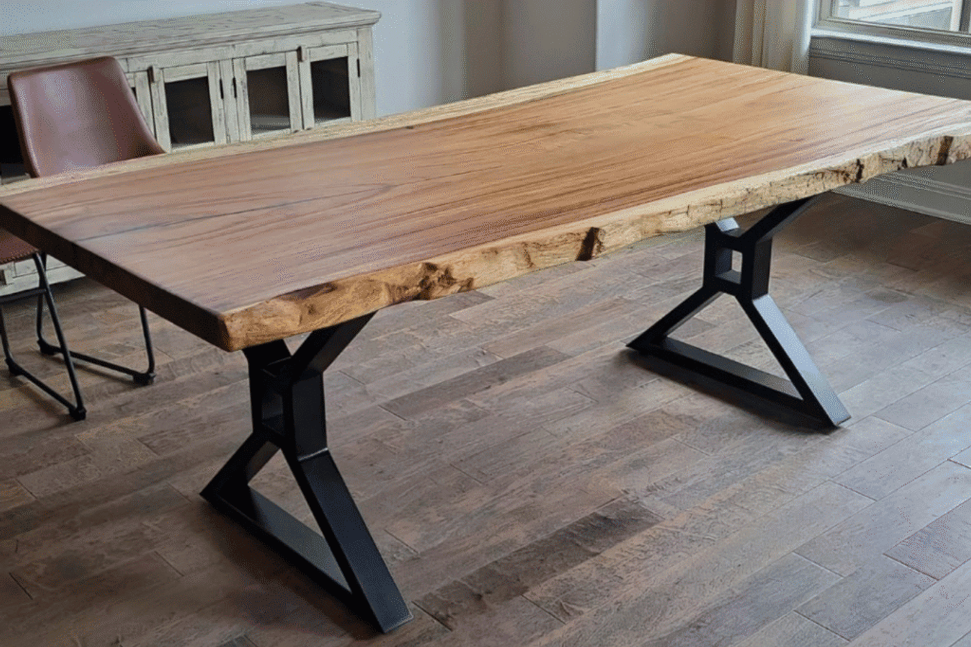 Live edge wooden dining table with natural wood grain and sturdy black metal legs, placed on a wooden floor in a bright room.