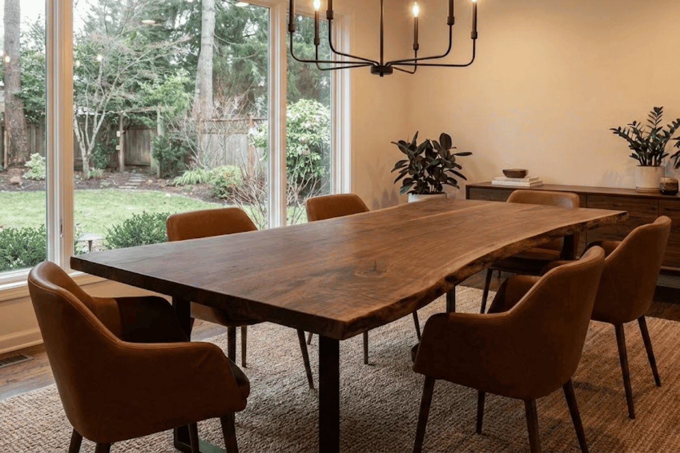 Dining room with live-edge wood table and six brown leather chairs beside large windows overlooking a green backyard