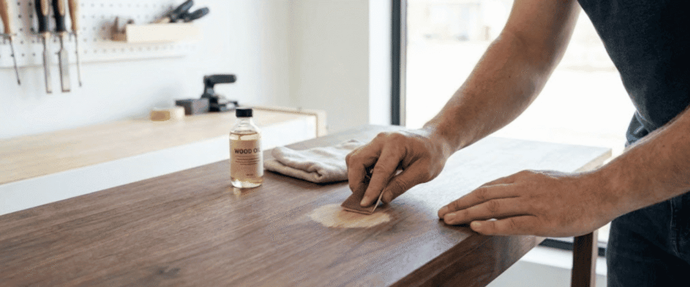 Person sanding and applying wood oil to a tabletop, hands smoothing a repaired patch with a cloth and small bottle of finish nearby