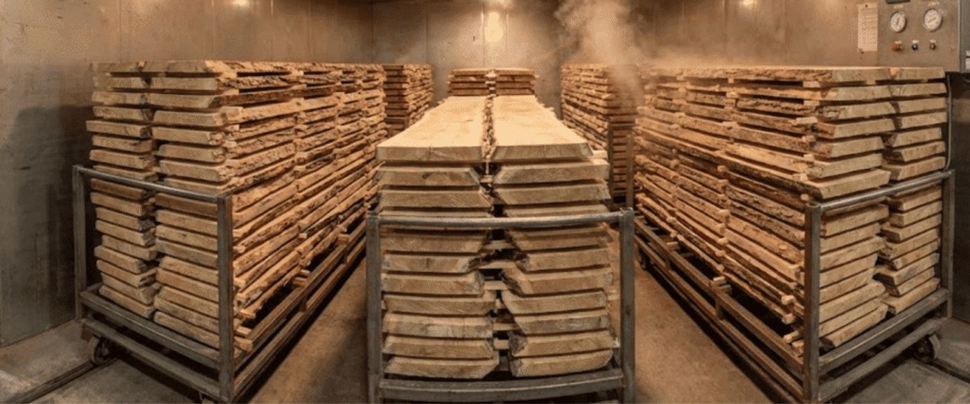 Stacked rough-cut oak boards on metal carts inside a climate-controlled kiln, steam rising as fans and controls regulate drying