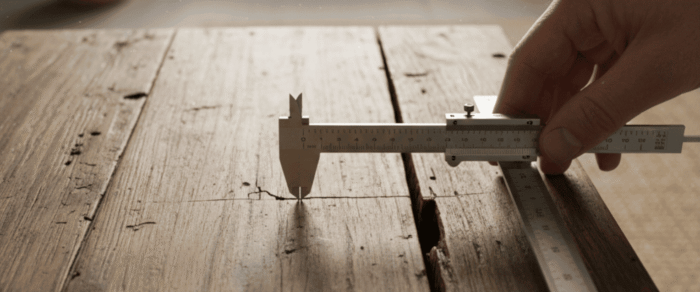 Hand holding a caliper measuring a crack gap on a weathered wooden table surface