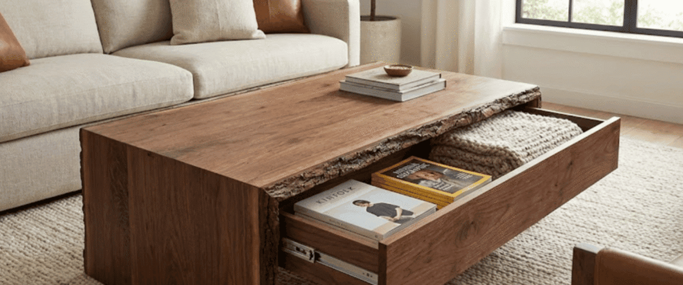Live-edge walnut coffee table with open drawer holding magazines and a knit blanket in a sunlit neutral living room