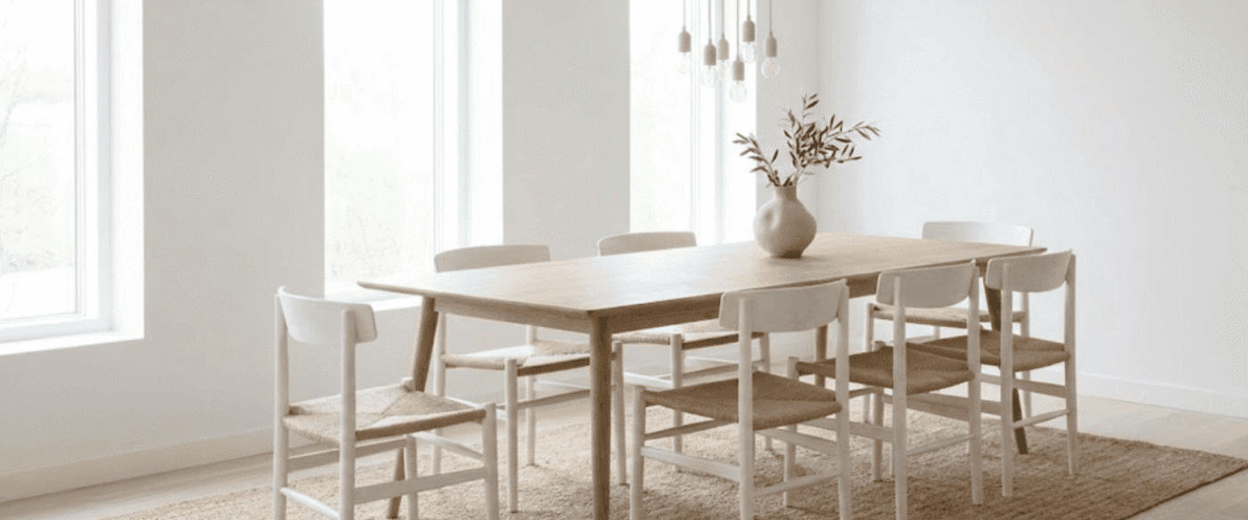 Calm minimalist dining room with light wood table, six woven-seat chairs, neutral rug and vase with greenery by bright windows