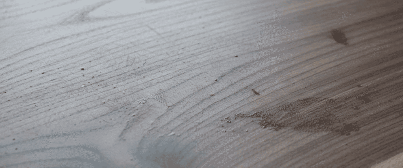 Dusty wooden tabletop with natural grain, small debris, a light water ring, and a knot near the edge
