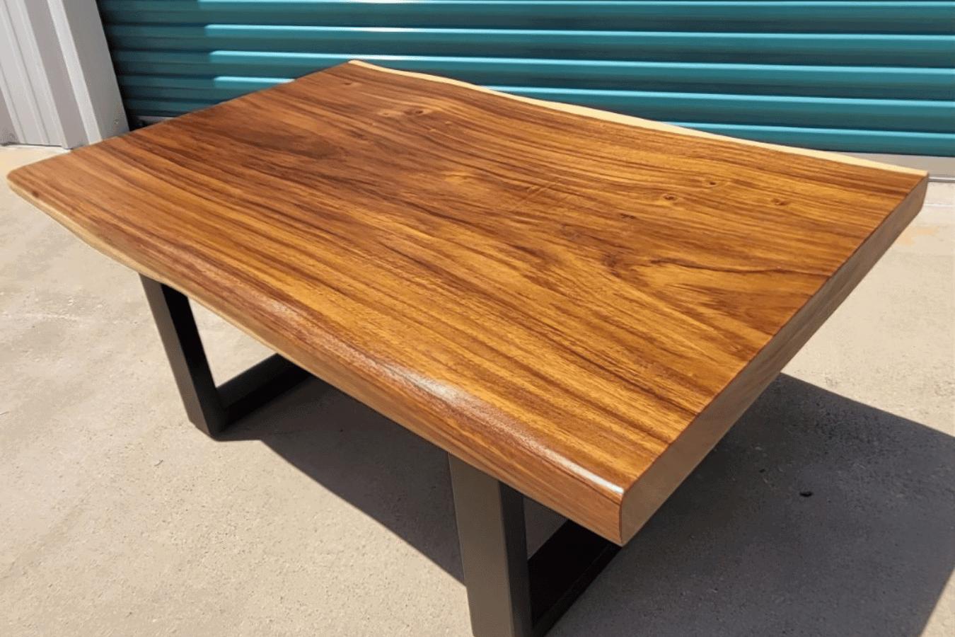 Polished live-edge wooden table with warm grain and black metal legs, photographed outdoors against teal storage doors.