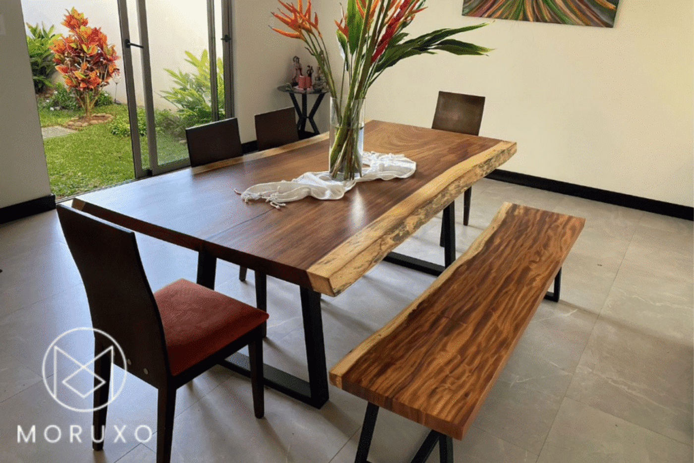 Natural wood dining table and bench with metal legs, paired with cushioned chairs. Modern, rustic style in a bright room with garden view.