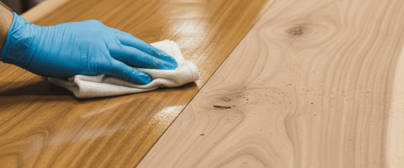 Gloved hand wiping wood tabletop with cloth, applying finish to bring out grain contrast between sealed and raw sections.