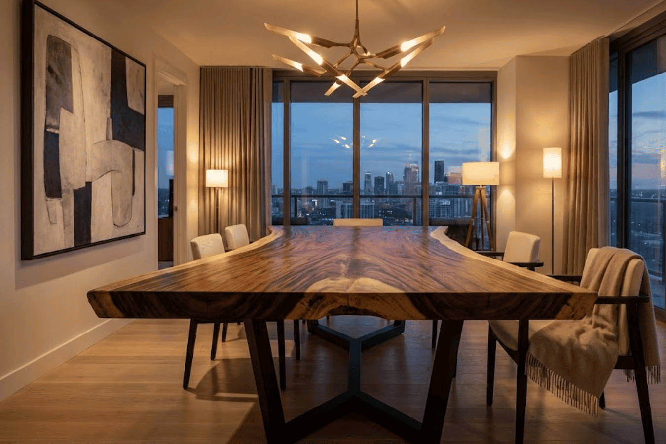 Dining room with a live-edge wooden table, modern chandelier, floor-to-ceiling windows showing city skyline at dusk, neutral chairs and warm lighting
