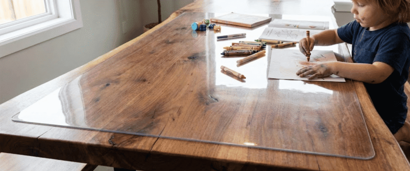 Child coloring at a wooden dining table with crayons and paper spread out, sunlight from a nearby window illuminating the scene