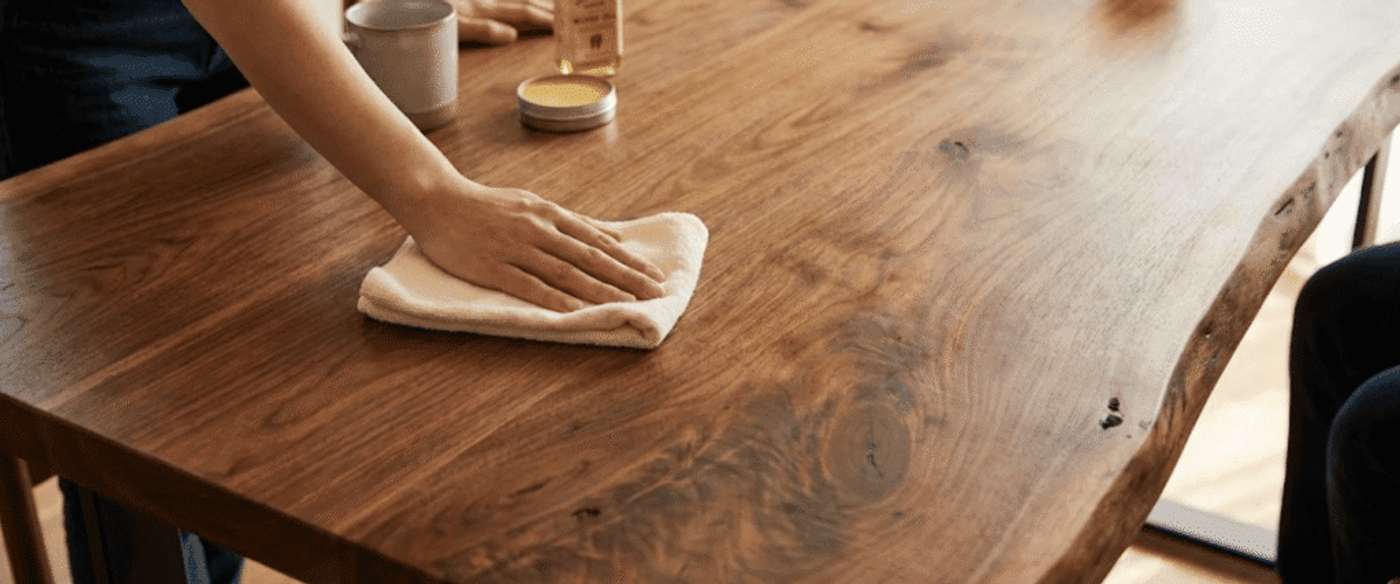 Person polishing a live-edge wooden table with a cloth, with wood oil and a mug nearby, hands visible