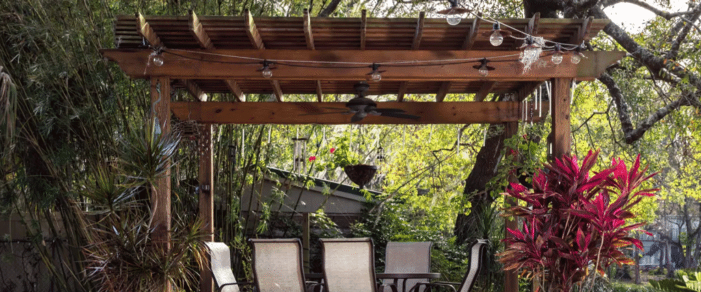 Wooden pergola with ceiling fan and hanging lights, outdoor dining chairs and table, surrounded by lush greenery and vibrant plants in a backyard garden