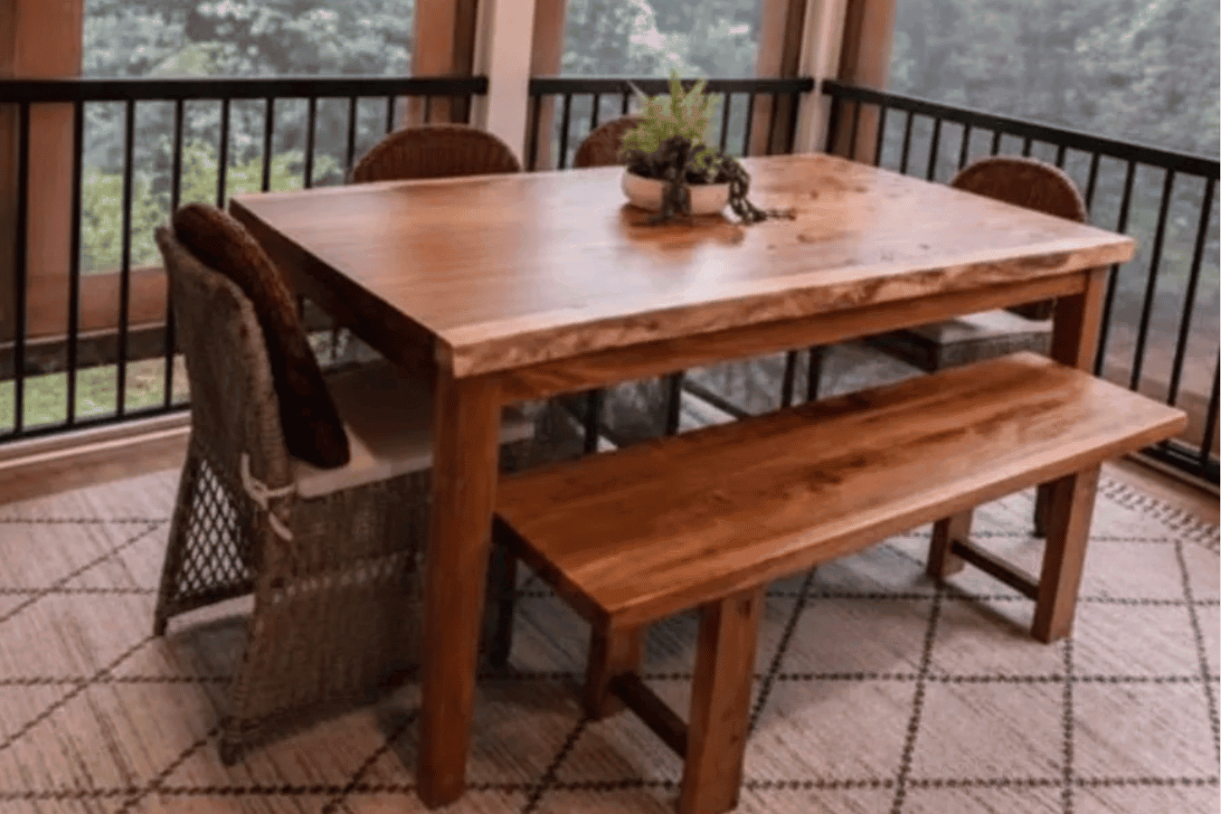 Sunlit wooden dining table and bench on a screened porch, surrounded by trees visible through large windows.