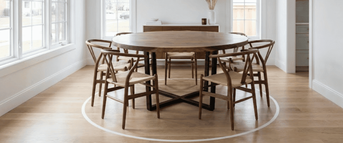 Round wooden dining table with six woven-seat chairs in a bright room, light hardwood floors and large windows