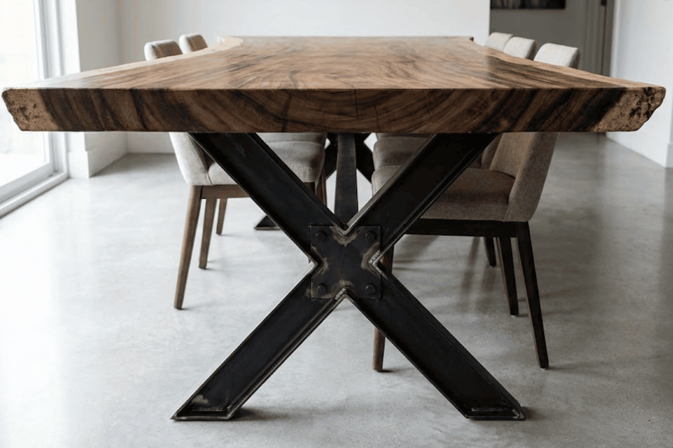 Rustic live-edge wood dining table with black metal X-shaped legs and upholstered chairs on a polished concrete floor