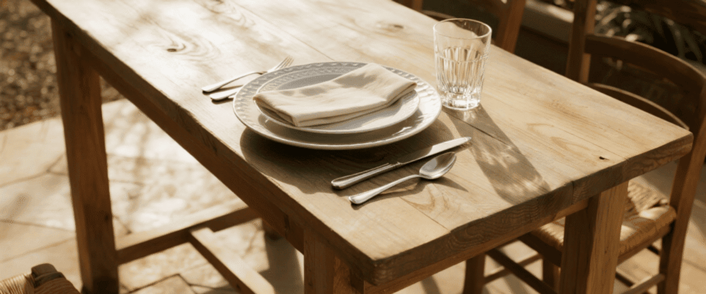 Sunlit wooden table set for one with a stacked plate, cloth napkin, fork, knife, spoon and a glass in a cozy outdoor dining spot.
