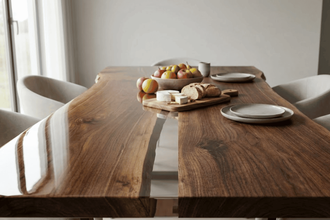 Polished live-edge wooden dining table with fruit bowl, bread and plates, surrounded by modern upholstered chairs in bright room