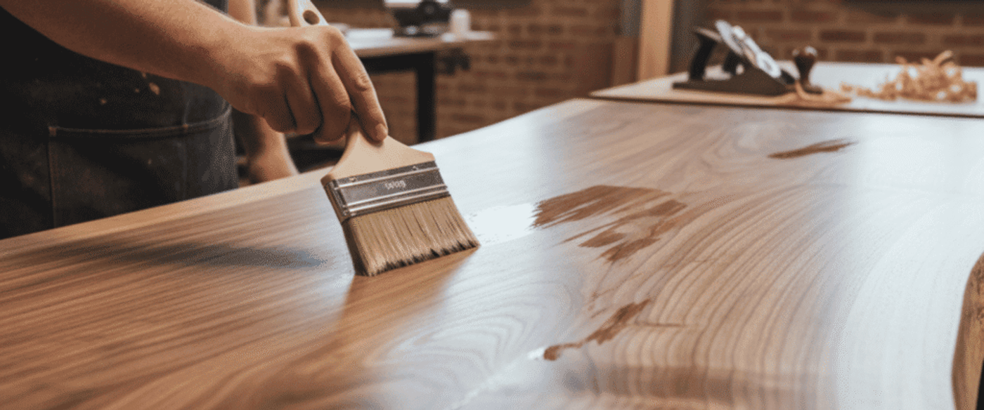 Woodworker applying clear finish with a brush to a live-edge walnut tabletop in a sunlit workshop, grain and sheen visible.