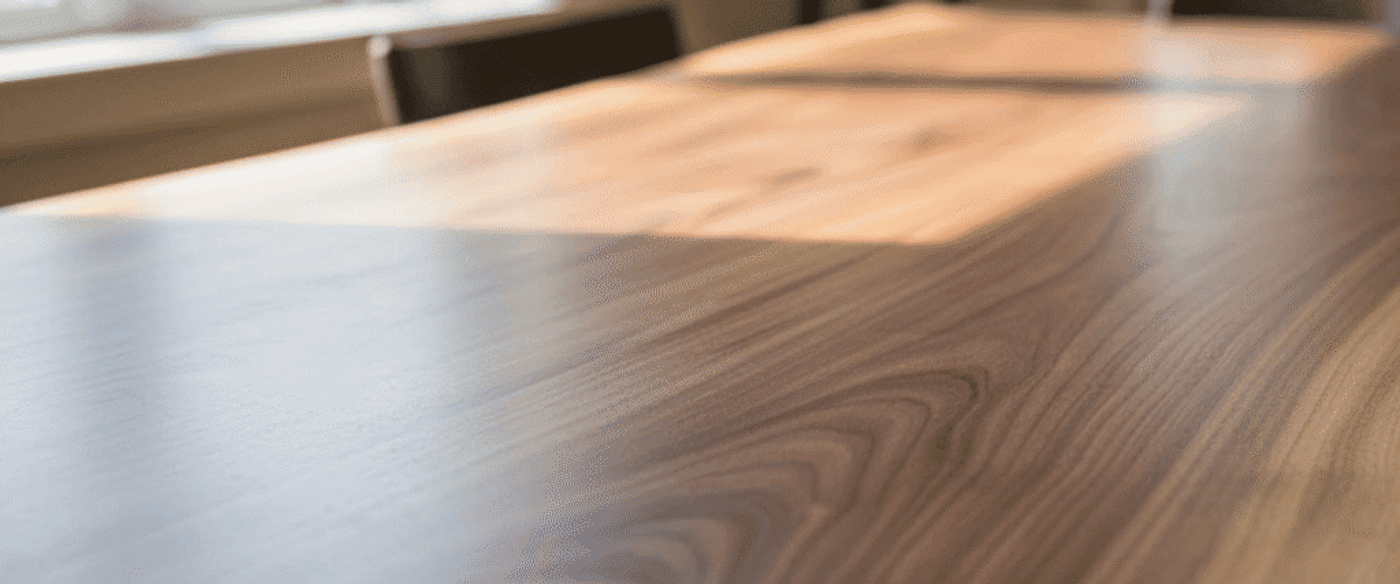 Sunlit wooden dining table with visible grain patterns, chairs and a lamp in a warm, minimalist room setting